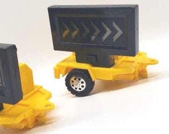 May include: Two miniature yellow construction trailers with black rectangular signboards. One sign displays a series of directional arrows, while the other features the partial text CAUTION! AHEAD!!!. Each trailer has a single black wheel.