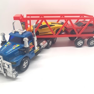 May include: A blue toy semi-truck with a silver front bumper and chrome exhaust stacks, pulling a red open-frame car carrier trailer. The trailer holds a yellow sports car and a red muscle car, both featuring black wheels and detailed body designs.