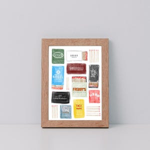Op de afbeelding: Een ingelijste print met een verzameling lucifersdoosjes met verschillende namen van restaurants en bedrijven. De lucifersdoosjes zijn in verschillende kleuren en ontwerpen, met zichtbare lucifers. De lijst is gemaakt van lichtbruin hout.