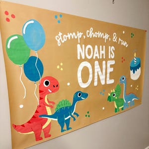 May include: A large banner with a brown background, featuring cartoon dinosaurs, balloons, and the text "stomp, chomp, & run NOAH IS ONE." The banner has illustrations of a red T-Rex, blue and green dinosaurs, and a birthday cake.