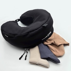 Könnte beinhalten: Ein schwarzes Reisekissen mit einer Reißverschlusstasche und dem Text "travel@day" an der Seite. Das Kissen ist geöffnet und enthält drei Paar Socken.