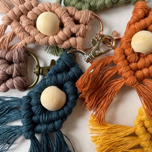 May include: A collection of handmade macrame keychains with wooden beads. The keychains are in various colors, including green, pink, orange, blue, and yellow. Each keychain has a unique design and features a wooden bead in the center.