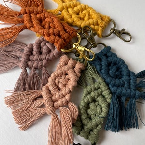May include: A set of six colorful macrame keychains with tassels. The keychains are made with different colored cords and feature a variety of knot patterns. Each keychain has a gold-toned metal clasp.