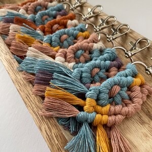 May include: A set of seven colorful macrame keychains with tassels. Each keychain features a geometric pattern in shades of blue, brown, pink, and yellow. The keychains are attached to silver key rings.