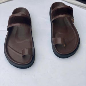 May include: A pair of brown leather sandals with a toe loop and a wide strap across the top of the foot. The sandals have a dark brown suede-like material on the top strap and a black sole. The sandals are designed for comfort and style.