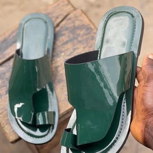 May include: A pair of green leather sandals with a glossy finish. The sandals have a toe loop and a wide strap across the top of the foot. The soles are white with a stitched border.