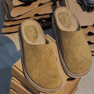 May include: A pair of tan suede slip-on clogs with a light-colored sole and a soft, cushioned interior. The clogs are displayed on a stack of cardboard boxes, suggesting a focus on footwear and comfort. A black belt is visible in the background.