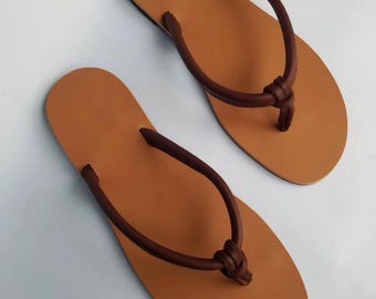 Op de afbeelding: Een paar bruine leren teenslippers. De sandalen hebben een lichtbruin voetbed en donkerbruine bandjes die bij de teen zijn geknoopt. De teenslippers staan tegen een witte achtergrond.