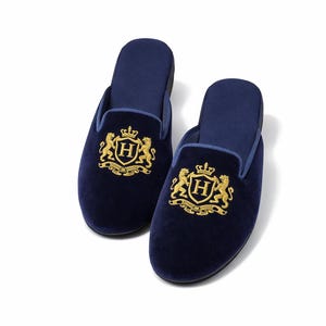 May include: A pair of navy blue velvet slippers. Each slipper has a gold embroidered crest with a crown, two lions, and the letter "H". The slippers have a slip-on design.