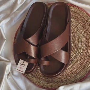 May include: A pair of brown sandals featuring wide crisscross straps and a toe loop design. The sandals have a smooth, solid-colored finish and a contoured footbed. A small white rectangular tag with black text is attached to the left sandal.