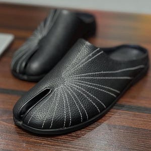 Pode incluir: Sapatos pretos de couro sem cadarços com costuras brancas. Os sapatos têm um design de bico dividido e são exibidos em uma superfície de madeira. As costuras irradiam da área dos dedos.