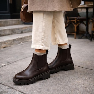 Könnte beinhalten: Dunkelbraune Chelsea-Stiefel mit einer klobigen, profilierten Sohle. Die Stiefel sind aus Leder gefertigt und haben elastische Seiteneinsätze. Sie werden mit cremefarbenen Hosen und grauen Socken getragen. Modische Schuhe.