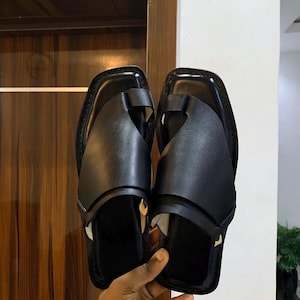 May include: A pair of black leather sandals with a square toe design. The sandals feature a strap across the top of the foot and a toe loop. The footwear is held up against a wooden door.