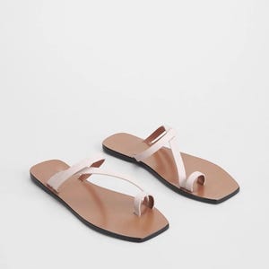 May include: A pair of light pink leather sandals with a toe loop and thin straps. The sandals have a brown footbed and a black sole. The sandals are displayed on a white background.