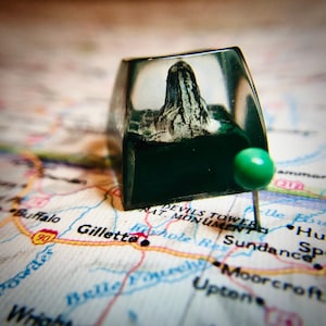 May include: A clear resin keycap with a miniature mountain inside. The keycap is sitting on a map with the text "Gillette" and "Devils Tower Nat. Monument" visible. A green push pin is stuck into the map.