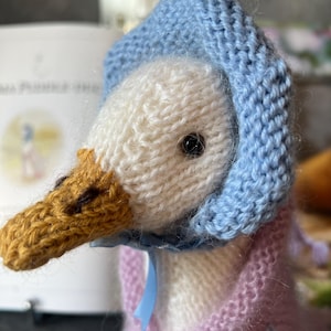 May include: A knitted goose doll wearing a blue knitted hat and a pink knitted dress with a blue ribbon. The doll has a brown knitted beak and black eyes.