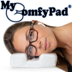 May include: A white pillow with a cutout for the eyes and nose. A woman is lying on the pillow with her eyes closed and wearing glasses. The pillow is labeled "My Comfy Pad" with a registered trademark symbol.