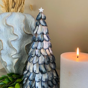 May include: A decorative Christmas tree made from blue and white seashells, topped with a white star. A lit white candle and a decorative vase with dried plants are in the background. A unique festive decoration.