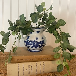 May include: A blue and white porcelain pitcher with a dragon design, filled with a green leafy plant, sits on top of two vintage books. The top book has the title "Battle-Fields of the Marne" and the bottom book has the title "Handbook of Business Letters".