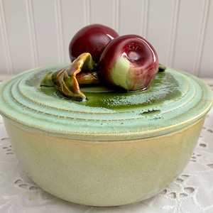 May include: A green ceramic bowl with a lid featuring two red apples and green leaves. The bowl has a light green glaze and a slightly raised rim.