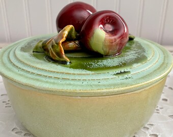 May include: A green ceramic bowl with a lid featuring two red apples and green leaves. The bowl has a light green glaze and a slightly raised rim.