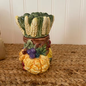 May include: A ceramic salt and pepper shaker set with a fall harvest theme. The shakers are shaped like a pumpkin and a bundle of wheat. The pumpkin is yellow with green leaves and a red stem. The wheat is green with brown stalks and yellow grains.