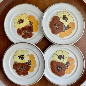 May include: Set of four white ceramic dessert plates with a floral design. The flowers are brown, yellow, and green.