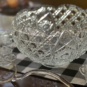 May include: A clear glass punch bowl with a cut glass design, a matching ladle, and several smaller glasses. The bowl and glasses are on a black and white checkered tablecloth.