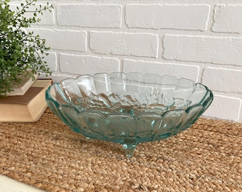 May include: A clear, teal-colored glass bowl with a scalloped rim and three small feet. The bowl features a grape design and is set on a woven mat. Decorative and functional.