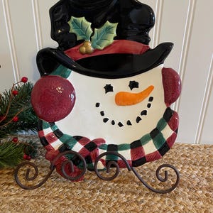 May include: A snowman-shaped decorative plate with a black top hat, red scarf, and a carrot nose. The plate has a checkered green, black, and red border and sits on a decorative metal stand. Festive holiday decor.