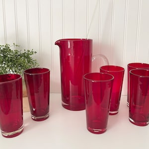 May include: Set of seven red glass tumblers and a matching pitcher. The glasses are tall and slender, while the pitcher has a wide base and a spout.