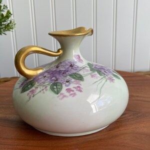May include: A white ceramic pitcher with a gold handle and a floral design of purple and green flowers. The pitcher is sitting on a wooden surface.