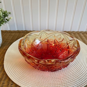 May include: A vintage, amber-colored glass candy dish with a lid. The dish has a starburst pattern and a scalloped edge. The dish is sitting on a white, woven placemat.