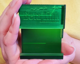 Puede incluir: Un dispositivo rectangular verde transparente con el texto "BetaLink Alpha Communicator" se sostiene en las manos de una persona. El dispositivo tiene un diseño en capas con una estructura interna visible. El fondo es de color amarillo liso.