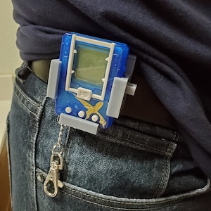 May include: A blue and grey digital pet toy with a screen and buttons. The toy is attached to a silver chain and is clipped to a pair of blue jeans.