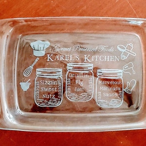 May include: A clear glass baking dish with two handles. The dish is engraved with the text "Gourmet Preserved Foods - Karel's Kitchen" and three jars labeled "Sizzlin' Sweet Nutz", "Drunkin' Fig Jam", and "Marvelous Marinara Sauce".