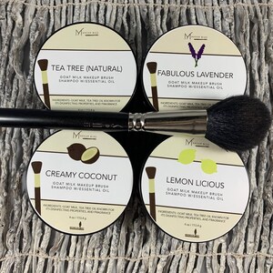 May include: Four round containers of goat milk makeup brush shampoo with essential oils. The containers are labeled with the scents: Tea Tree (Natural), Fabulous Lavender, Creamy Coconut, and Lemon Licious. A black makeup brush is laying on top of the containers.