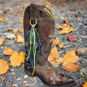 May include: A brown leather cowboy boot with floral embroidery and a decorative feather charm. The charm features blue, yellow, and green accents, along with a leopard print design. The text "Holoholo Custom Designs" is visible on the boot.