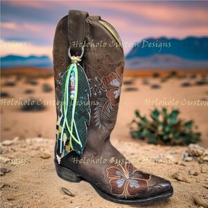 May include: Brown leather cowboy boot with floral embroidery and a decorative feather charm. The boot features a pointed toe and a stacked heel. The charm includes beads and ribbons in various colors. The text "Holoholo Custom Designs" is visible.