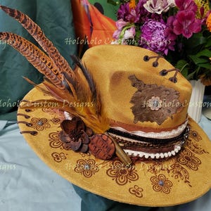 May include: A mustard-colored hat with brown floral patterns and decorative feathers. The hat features a brown leather patch, a decorative band, and a flower embellishment. The brim is adorned with brown floral designs and small jewels.
