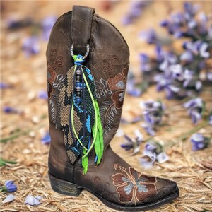 May include: Brown leather cowboy boot with floral embroidery and a decorative boot charm. The charm features a snakeskin-patterned feather, blue and green accents, and beaded details, adding a touch of flair to the Western-style boot.