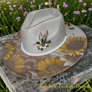May include: A light gray fedora hat featuring a large, colorful hummingbird and flower brooch on the front, a white cord trim around the base, and a wide brim decorated with a printed pattern of yellow sunflowers and small butterflies.
