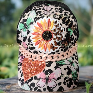 May include: A patterned trucker cap with a black mesh back, featuring a sunflower, butterflies, and a glitter heart. The cap has a pink chain detail and a black brim.