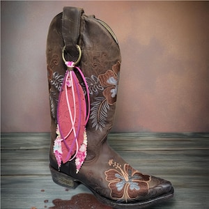 May include: A brown leather cowboy boot with floral embroidery and a decorative tassel. The tassel features a pink, red, and white feather design, attached to a metal ring. The boot is decorated with brown and blue floral embroidery.