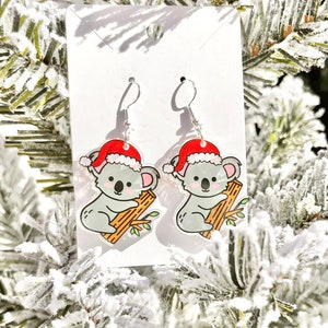 May include: A pair of Christmas-themed earrings featuring a koala wearing a Santa hat and holding onto a branch. The earrings are made of acrylic and have silver hooks.