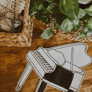 Pu&ograve; includere: Un ritaglio a forma di pianoforte in bianco e nero su una superficie di legno. Il pianoforte ha un corpo e gambe nere, con tasti bianchi. Un cestino intrecciato con strumenti musicali in legno &egrave; sullo sfondo. Sono visibili anche piante verdi.