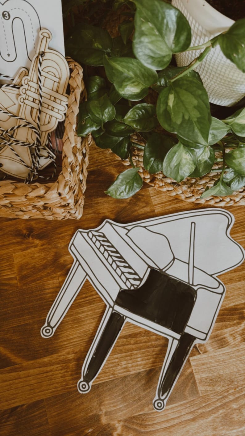 Pu&ograve; includere: Un ritaglio a forma di pianoforte in bianco e nero su una superficie di legno. Il pianoforte ha un corpo e gambe nere, con tasti bianchi. Un cestino intrecciato con strumenti musicali in legno &egrave; sullo sfondo. Sono visibili anche piante verdi.