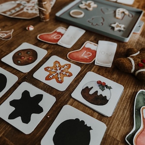 Puede incluir: Una mesa de madera exhibe artículos con temática navideña. Hay galletas de jengibre, una bandeja para hornear con formas de galletas y tarjetas con ilustraciones festivas. También hay tarjetas de calcetines rojos con números y un hombre de jengibre, creando una escena navideña.