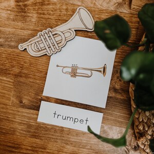 Pu&ograve; includere: Sagoma di tromba in legno e una carta con un'illustrazione di tromba e la parola "trumpet". L'illustrazione della tromba &egrave; color oro. La sagoma in legno &egrave; marrone chiaro. La carta &egrave; bianca. Lo sfondo &egrave; una superficie di legno.