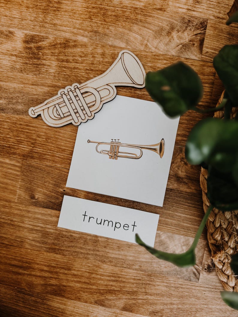 Pu&ograve; includere: Sagoma di tromba in legno e una carta con un'illustrazione di tromba e la parola "trumpet". L'illustrazione della tromba &egrave; color oro. La sagoma in legno &egrave; marrone chiaro. La carta &egrave; bianca. Lo sfondo &egrave; una superficie di legno.